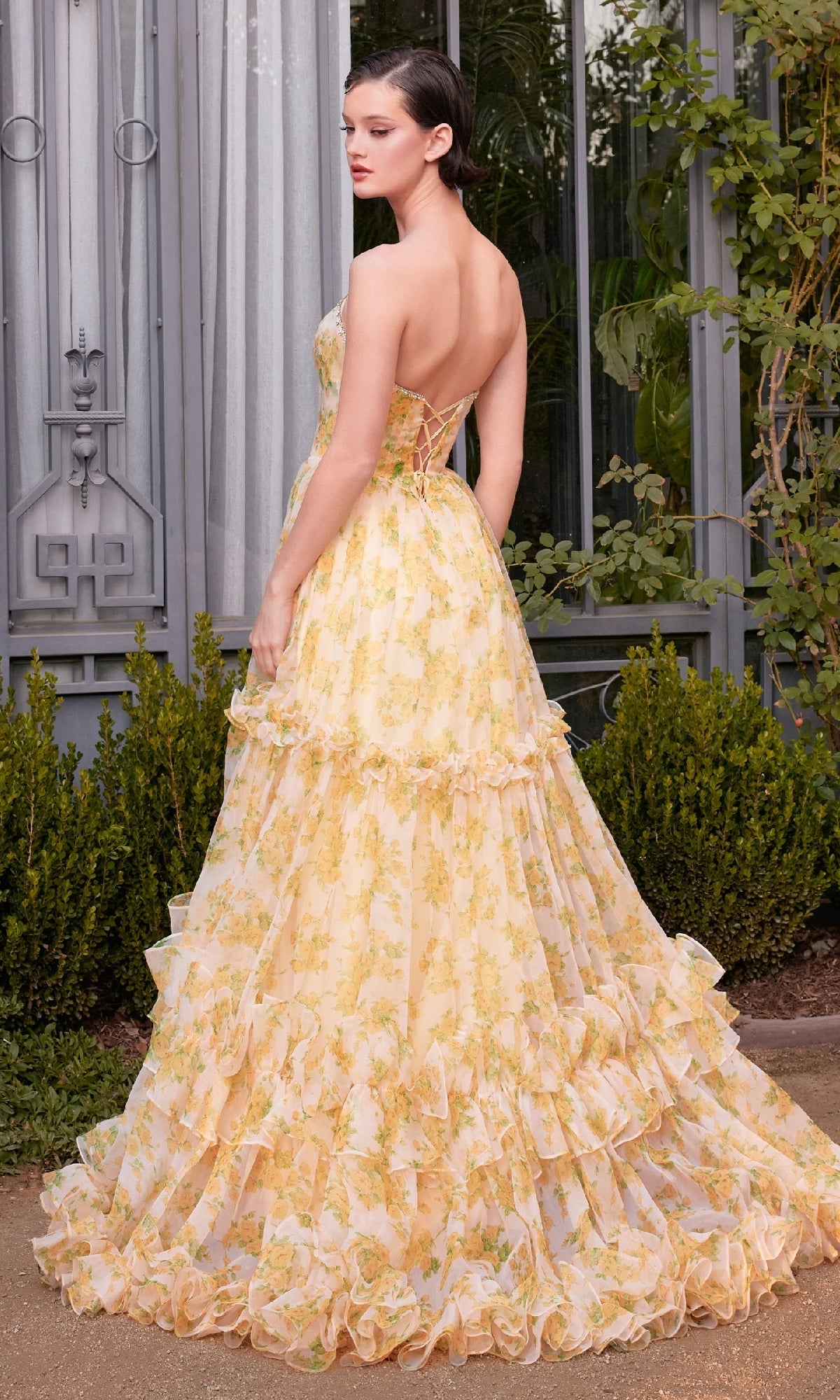 A woman stands outdoors near green bushes and a window, wearing the Andrea & Leo A1430 Yellow Flora-Print Prom Gown, featuring a strapless design, ruffled layers, and a ball gown silhouette.